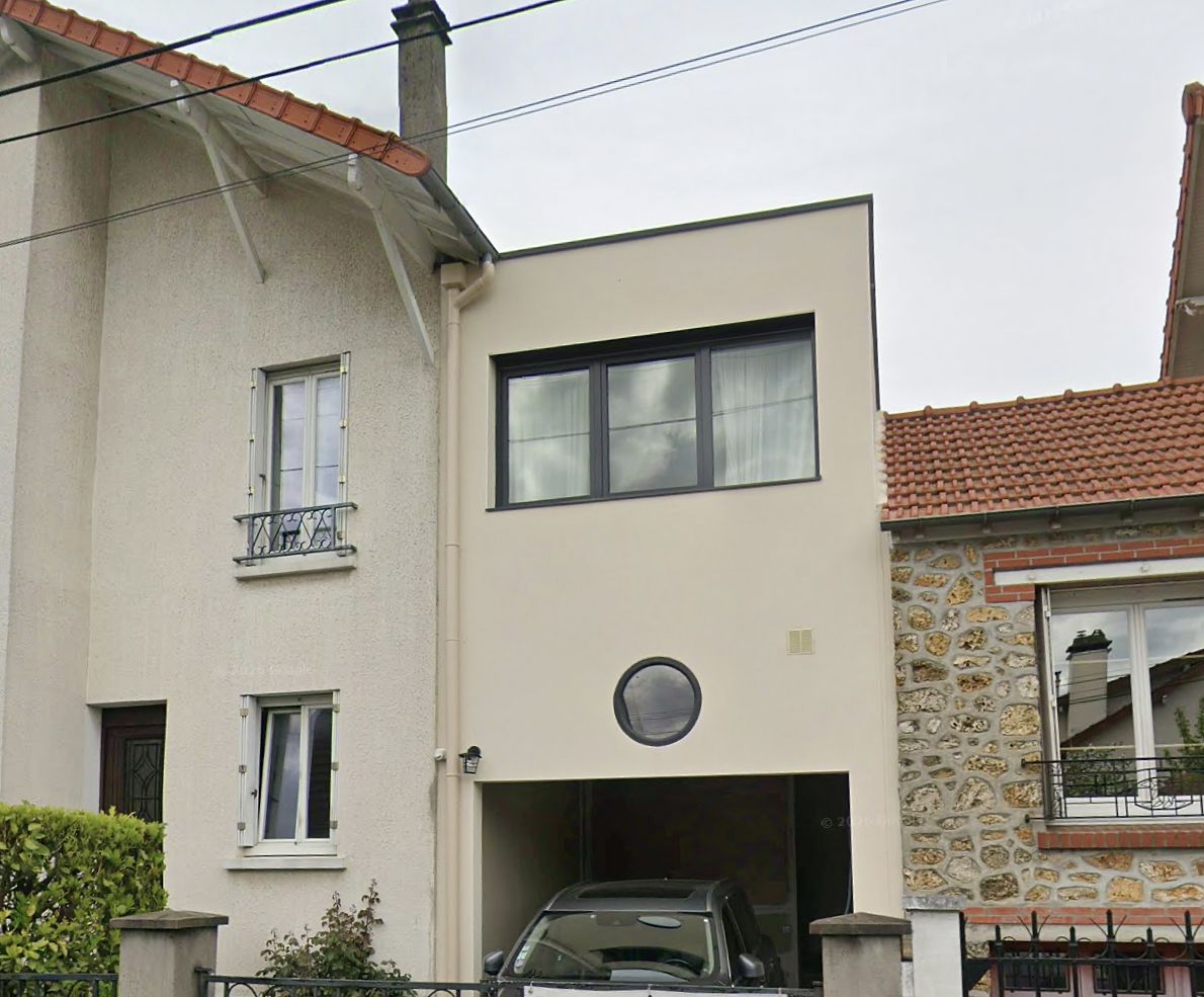 Extension construite au-dessus du garage d'un pavillon, enduit clair, grande baie horizontale et œil-de-bœuf